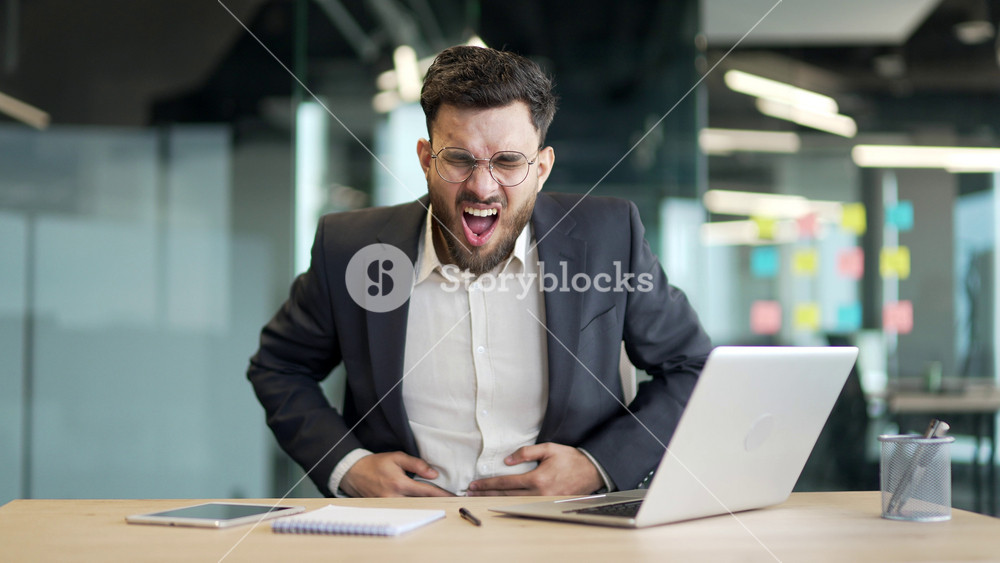 Sick businessman in a formal suit feels stomach pain working on laptop sitting at workplace in business office. Worker has heartburn, gastritis or poisoning. Male suffers from spasms and constipation
