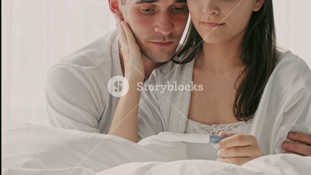 In a bright bedroom, a young couple sits together on the bed, holding a pregnancy test with a mix of hope and anticipation. Their close