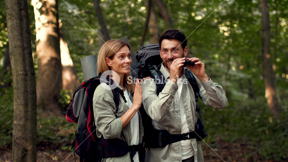 A woman with light skin and long hair stands beside a man with dark hair as he uses binoculars to observe nature. They have backpacks and