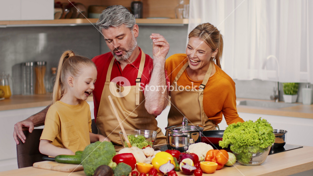 Smiling Adult Caucasian Male And Female Cooking Together With Young Girl In Bright, Modern Kitchen. Family Bonding While Preparing Healthy
