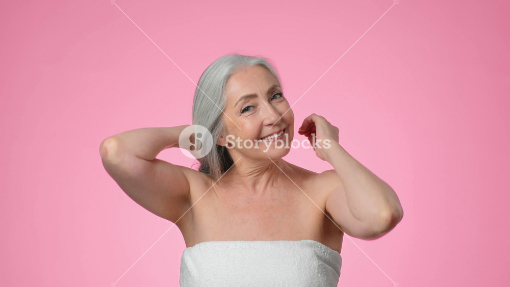 A woman with gray hair stands against a pink backdrop, smiling and touching her hair. She exudes confidence and joy in this moment. The