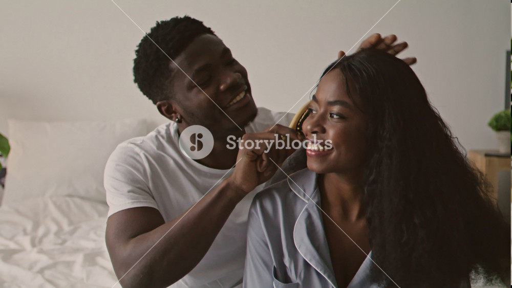 A man with dark skin is grooming the hair of a smiling Black woman in a relaxed setting at home. Soft light fills the room as they share a