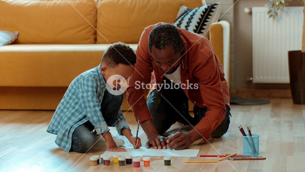 A man with dark skin and a boy are sitting on the floor of a living room. They are focused on drawing and painting on a sheet of paper. Art