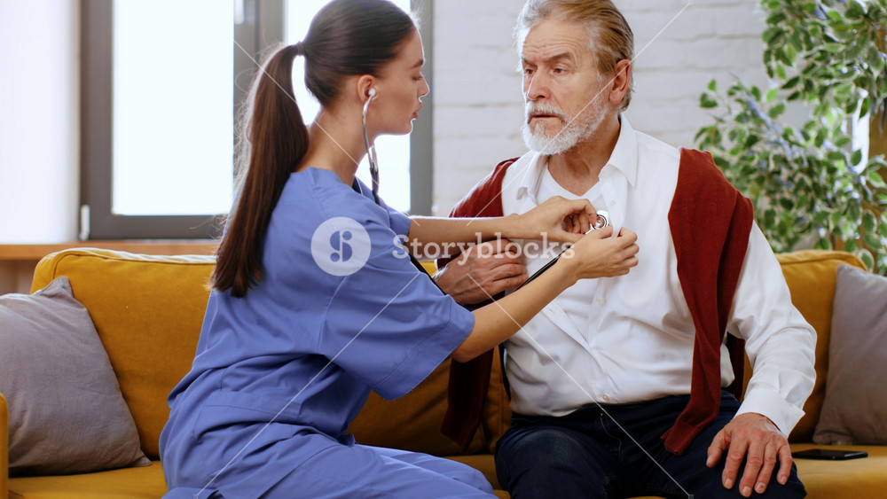 A woman in blue scrubs uses a stethoscope to check the heartbeat of an older man with gray hair and a white shirt, while he sits comfortably