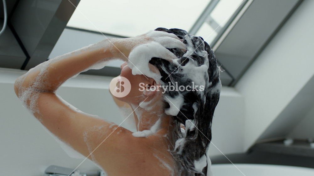 A woman is in a bright and airy bathroom, washing her hair with shampoo. She smiles as she enjoys the soothing water and rich lather. The