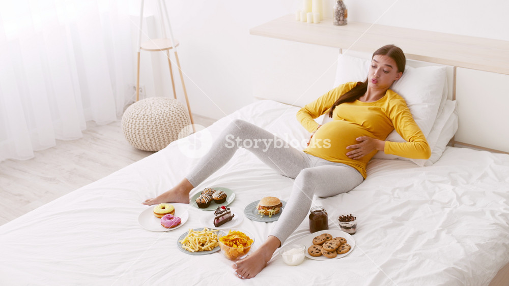 A pregnant woman sits comfortably on a bed dressed in casual attire, feeling full, surrounded by an of junk food including burgers, donuts