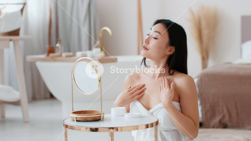 After shower bodycare. Young asian woman wrapped in towel applying pampering lotion on shoulders and neck, moisturizing body at bathroom