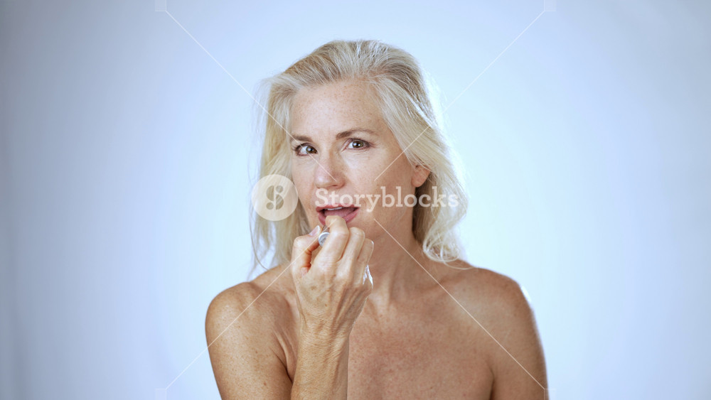 Portrait of mature middle-aged elderly woman makeup hygienic lipstick applying moisturizing lip balm natural beauty face skin care anti-aging cosmetic senior lady smiling light blue studio background