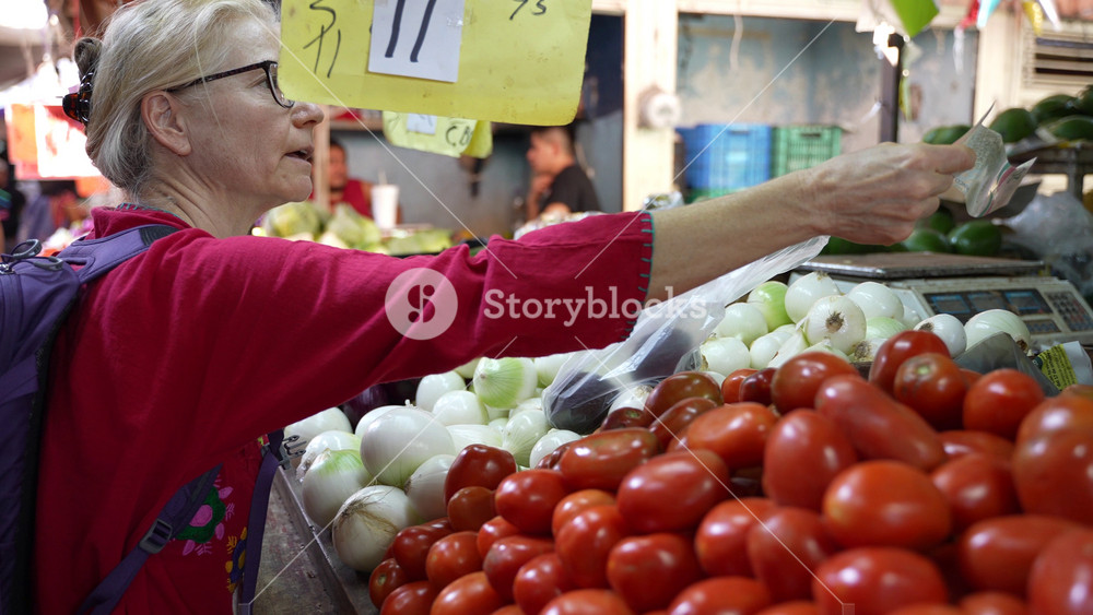 Pretty blonde mature senior woman wearing ethnic attire, exchanging money for buying vegetables and fruit at a local indoor market in Mexico.