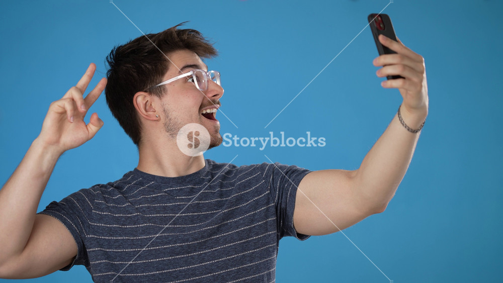 Fun young brunet man 20s wears striped t-shirt purple shirt doing selfie shot on mobile cell phone post photo on social network meet greet wave hand isolated on blue background studio portrait