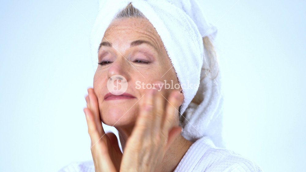 Closeup portrait of happy senior european woman applying moisturizer with fingers, looking off camera and smiling, female doing makeup wearing robe and towel on head