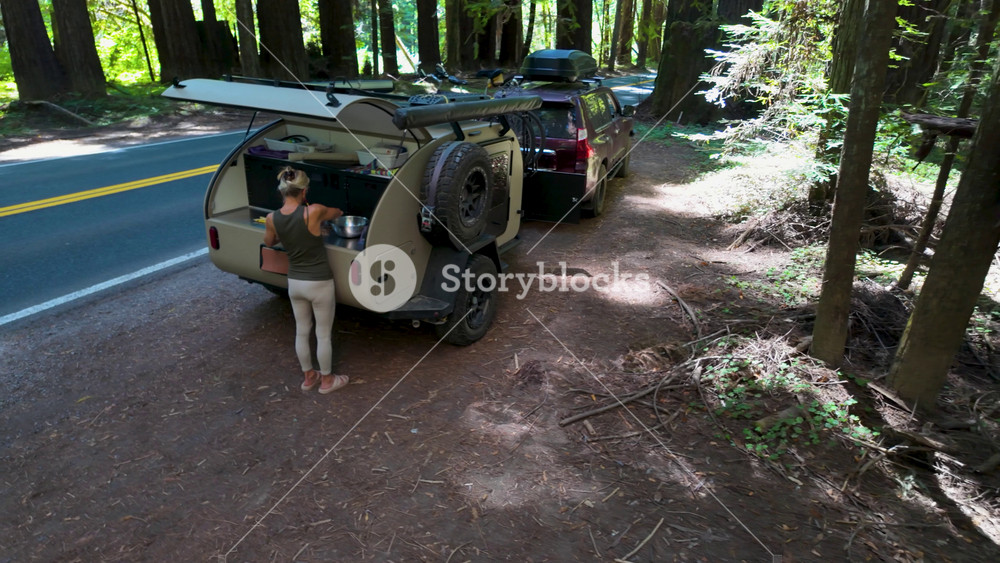 A woman prepares lunch at a teardrop camper parked along the road in Navarro River Redwood State Park. Beautiful redwood trees surround her