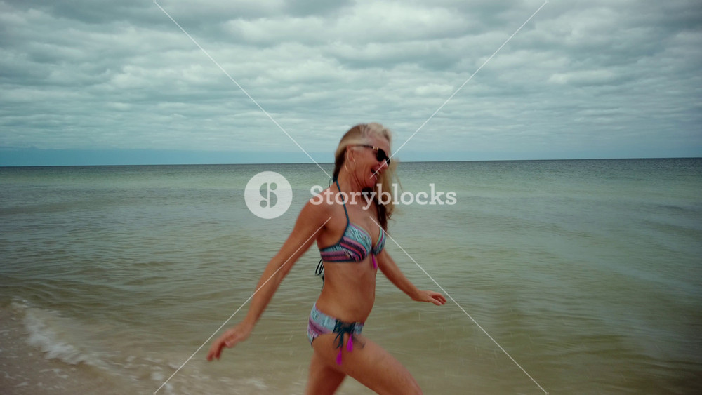 Side shot of playful blonde mature woman in sunglasses and bikini running along the shore laughing and putting her arms up in the air showing joy.