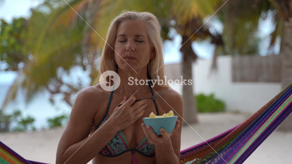 Closeup of pretty mature woman in bikini sitting in hammock at a resort, smells a piece of pineapple and then tastes it and smiles at the camera.