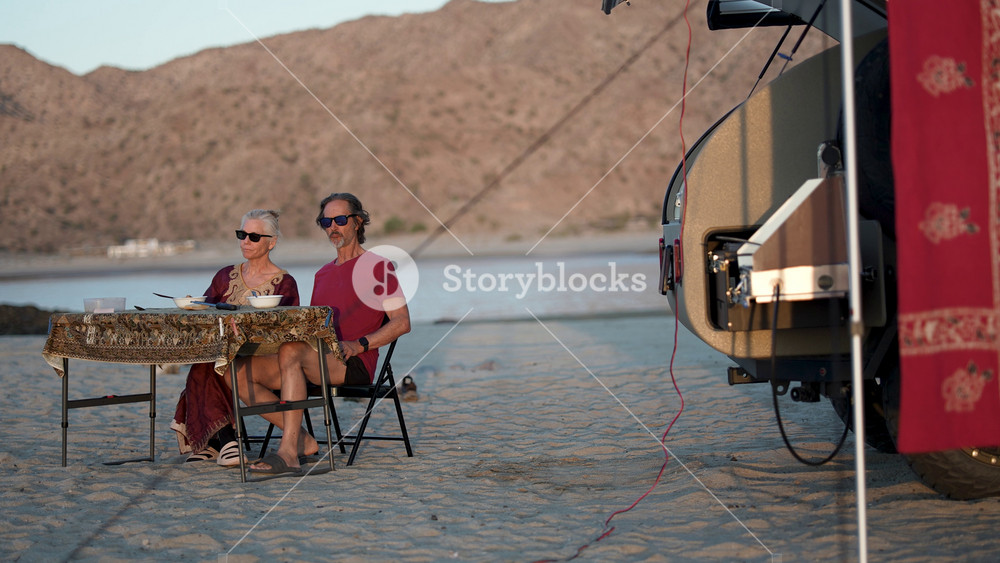 Mature man and woman dine outside their teardrop camper on a beach in Baja California as the sun sets over the water