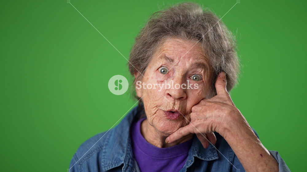 Closeup slow motion funny portrait of smiling happy elderly senior old woman with wrinkled skin and grey hair smiling and giving call me gesture with hands isolated on green screen background.