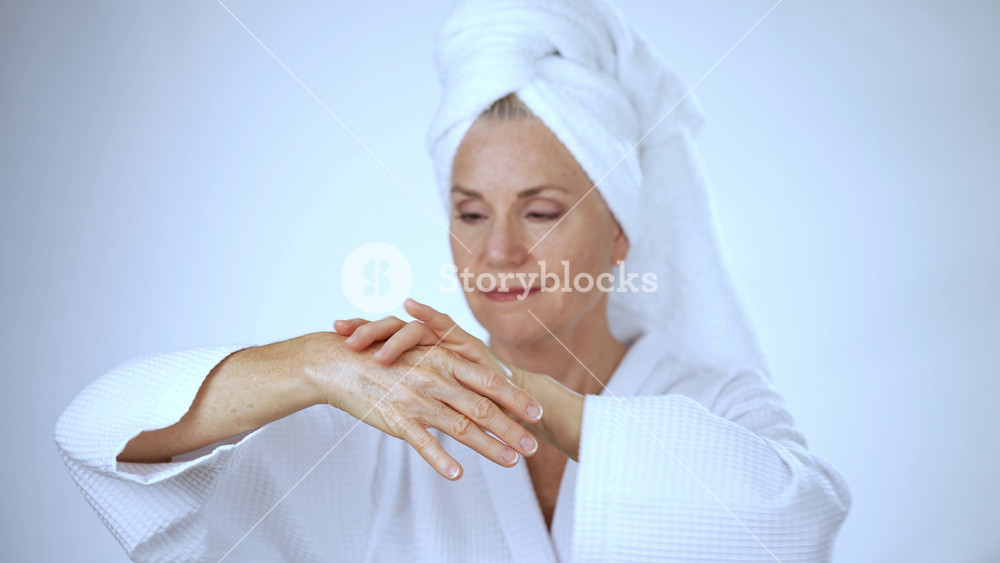 Middle-aged mature woman wearing robe and towel apply cream on hand, white background female hands care rub moisturizing body lotion daily beauty treatment soft skin anti-aging cosmetics rejuvenation