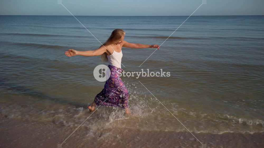Slow motion of mature woman in sarong splashing on the shore of a beach and turning around with arms outstretched.