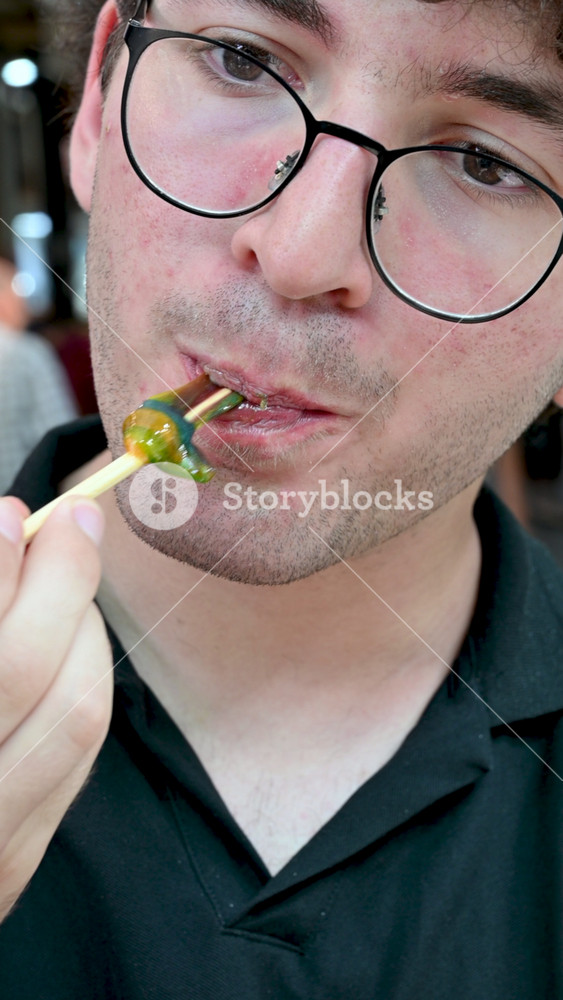 Izmir,Turkey,august 8, 2025.Young man with curly hair and glasses eating a colorful and sticky traditional ottoman candy, known as macun, on a wooden stick.He is savoring the sweet and sugary taste.