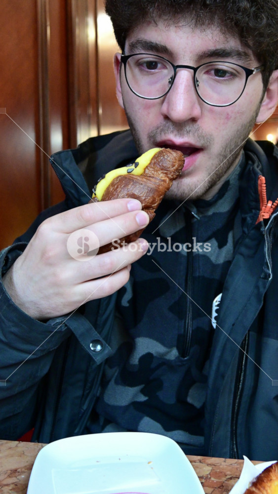Caucasian man with glasses eating a delicious italian croissant filled with custard at the bar. Morning routine in a typical cafe, a moment of pleasure enjoying a sweet pastry with pleasure
