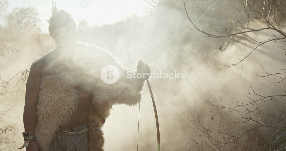 Male barbarian with bow standing in fume and looking away during hunt in wilderness against the background of smoky forest. Preparation for