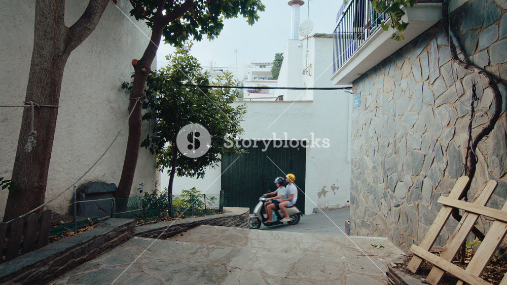Small mediterranean town alleyway or street, with white cobble stones and old houses. Tourists and locals on retro vintage mopeds drive up