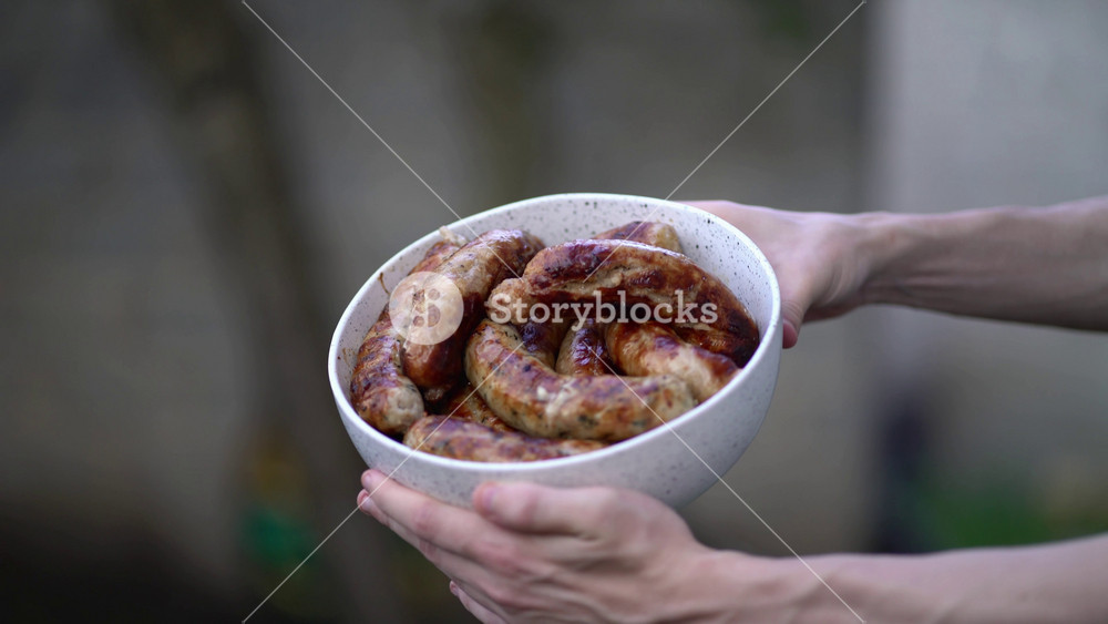 Golden brown grilled sausages with char marks in speckled bowl held by hands. Appetizing bbq food perfect for summer picnics and outdoor cooking videos.