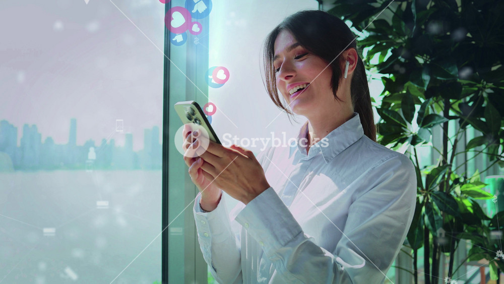 Asian woman wearing wireless earphones while standing near window. Pretty female holding smartphone with holographic social media icons