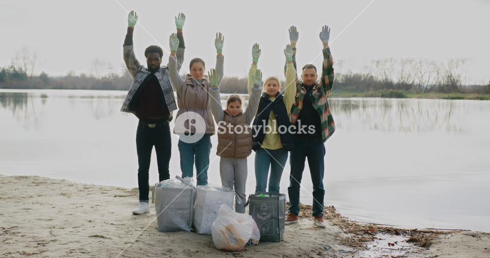 Camera view of group of people standing in front of big lake at beach. Everyone looking directly. Raising their hands up. Volunteers