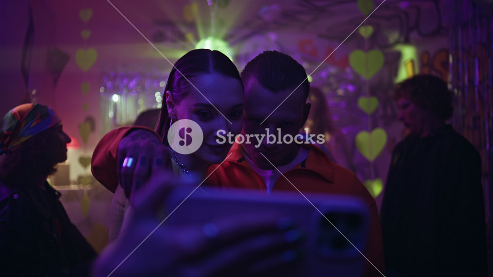 Clubbing couple taking selfie under neon lights at club night closeup. Trendy friends photographing on smartphone standing vivid dance floor