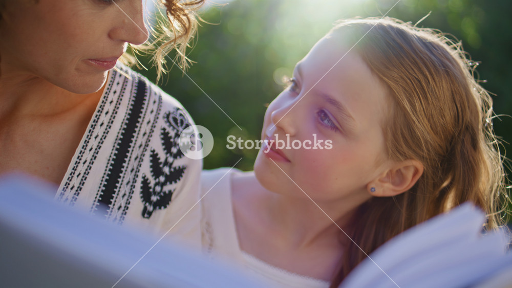 Mom kid reading book sitting nature closeup. Adorable small girl talking with woman parent spending weekend at summer park. Smiling mother