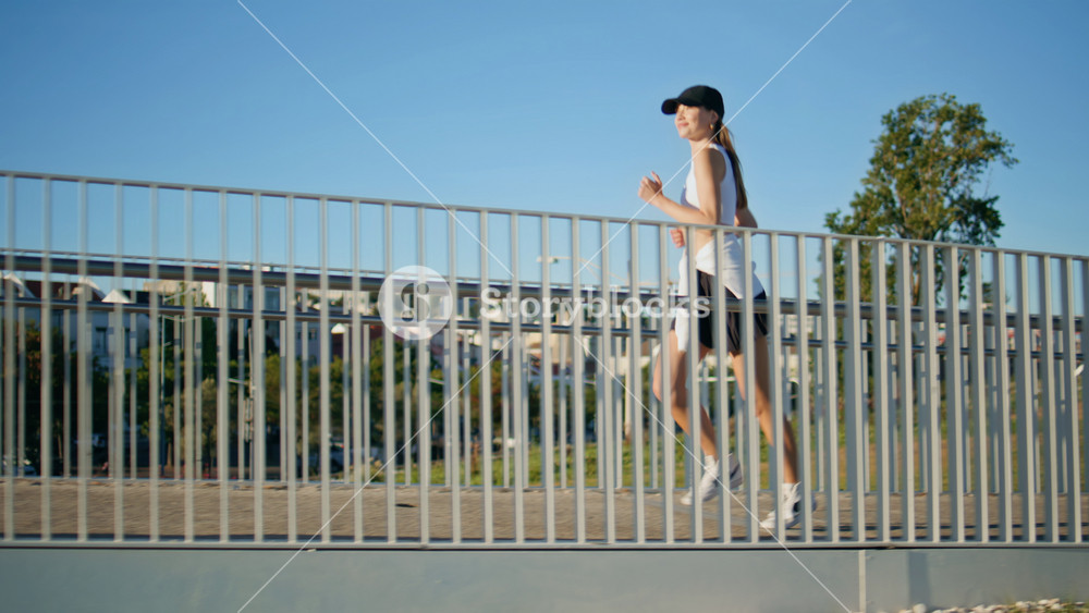Athletic woman running bridge in front clear blue sky. Energetic fitness lady jogging sunny city street wearing black cap. Enthusiastic