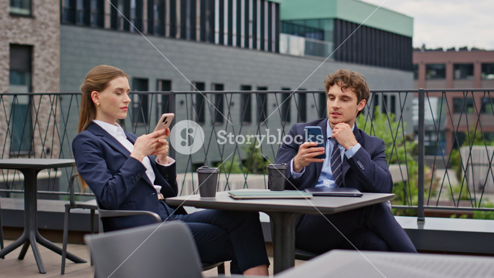 Businessman businesswoman relaxing at street cafeteria together. Smiling colleagues in suits enjoying casual break holding smartphones