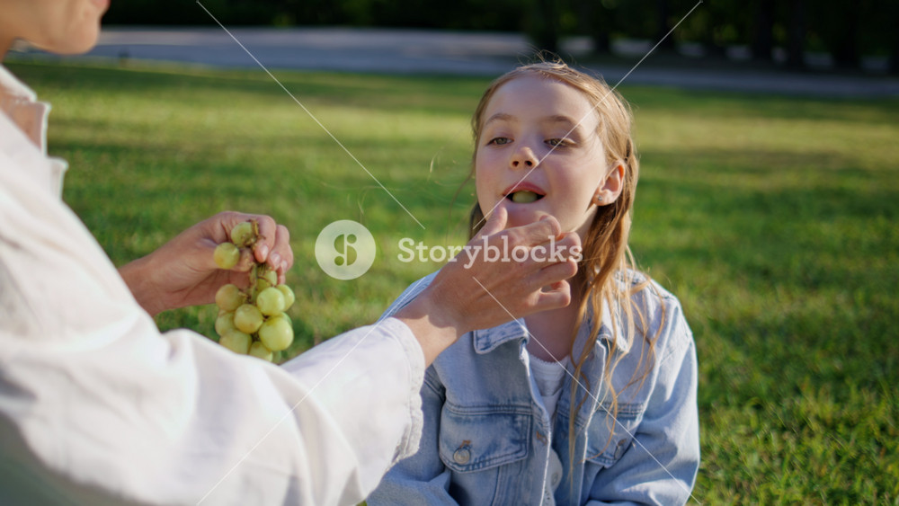 Attractive mother giving to daughter sitting at green closeup. Unrecognizable mom propose tasty food to blond girl on weekend. Young family