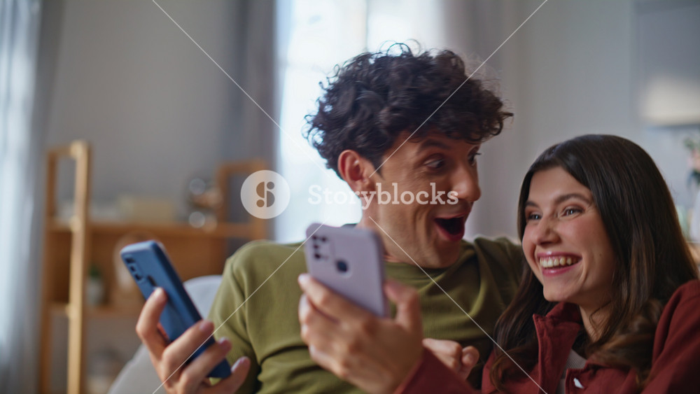 Amazed winners watching smartphones reacting success at living room together closeup. Overjoyed soulmates celebrating winning lottery
