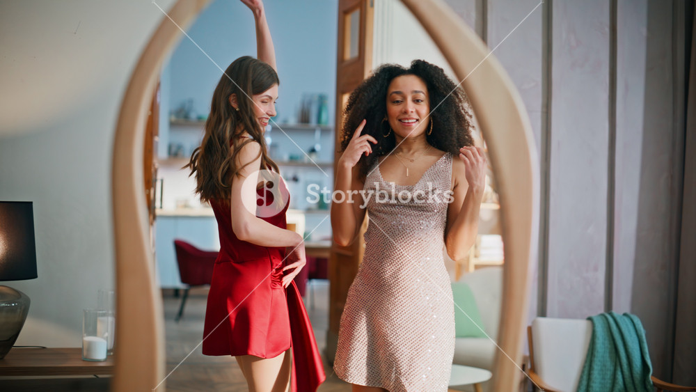 Gorgeous girls dancing mirror enjoying fashionable dresses. Happy two multiracial women admiring classy outfits moving bodies. Confident
