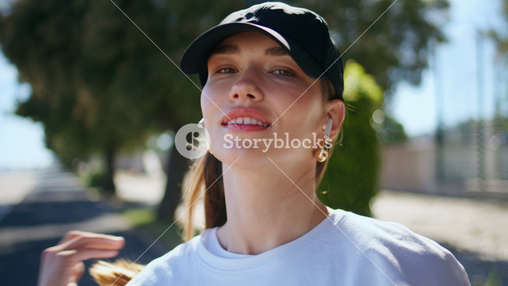 Happy girl adjusting hair smiling to camera at sunny city street closeup. Portrait relaxed woman wearing cap listening music in wireless