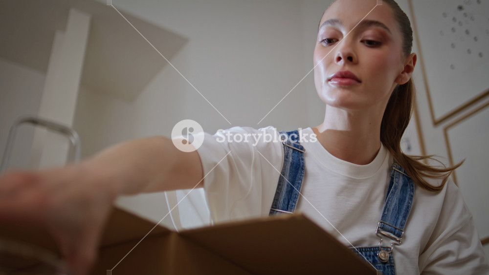 Woman unboxing household items moving in new apartment closeup. Young lady unpacking cardboard box with vase in home living room. Casual