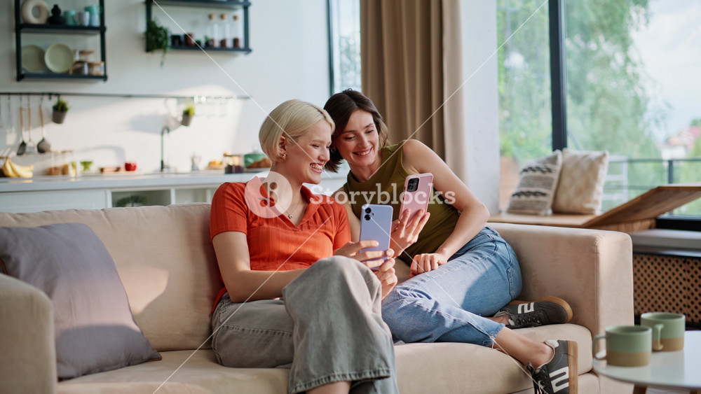 Relaxed sisters looking smartphones in modern house. Laughing girls friends using mobile phones on sofa in living room. Happy women having