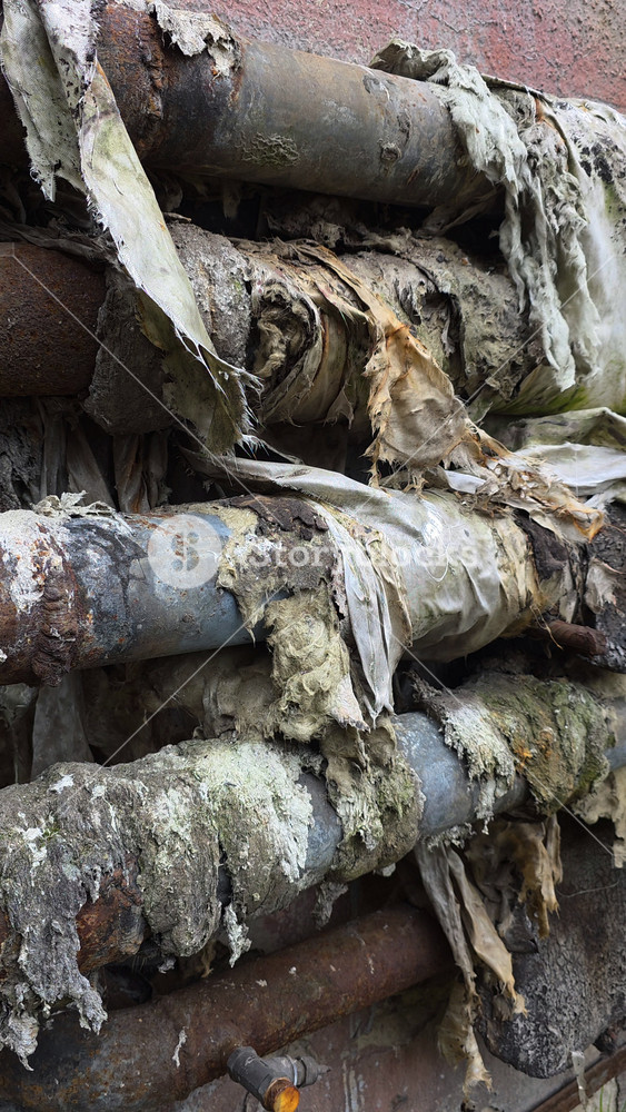 Damaged and peeling insulation on old rusty industrial heat pipes with corrosion, mold, and exposed fiberglass, illustrating neglect, urban