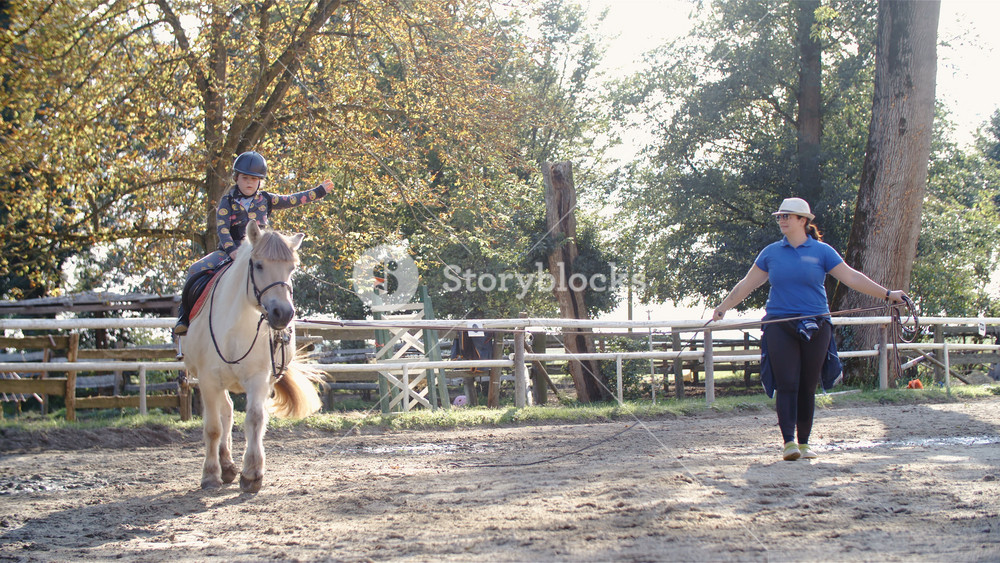 Teaching gymnastics while riding horse in slow motion 4K. Wide shot of child and trainer in focus training in the middle of outdoor riding arena on a sunny day. Horse on lounge walking in a circle.