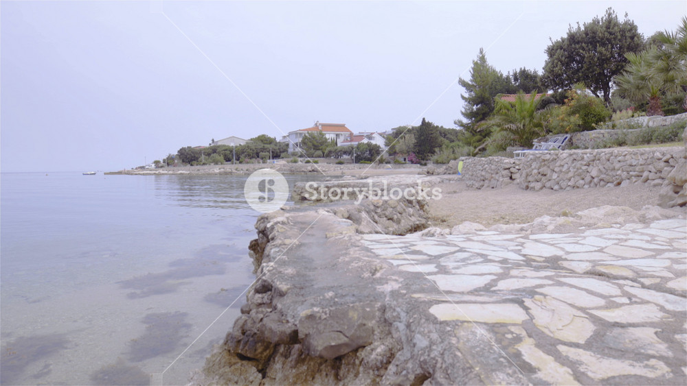 Empty stone paved beach dolly slide 4K. Wide shot of sea and beach in focus sliding towards the sea while no person around. Trees and apartments in background out of focus.