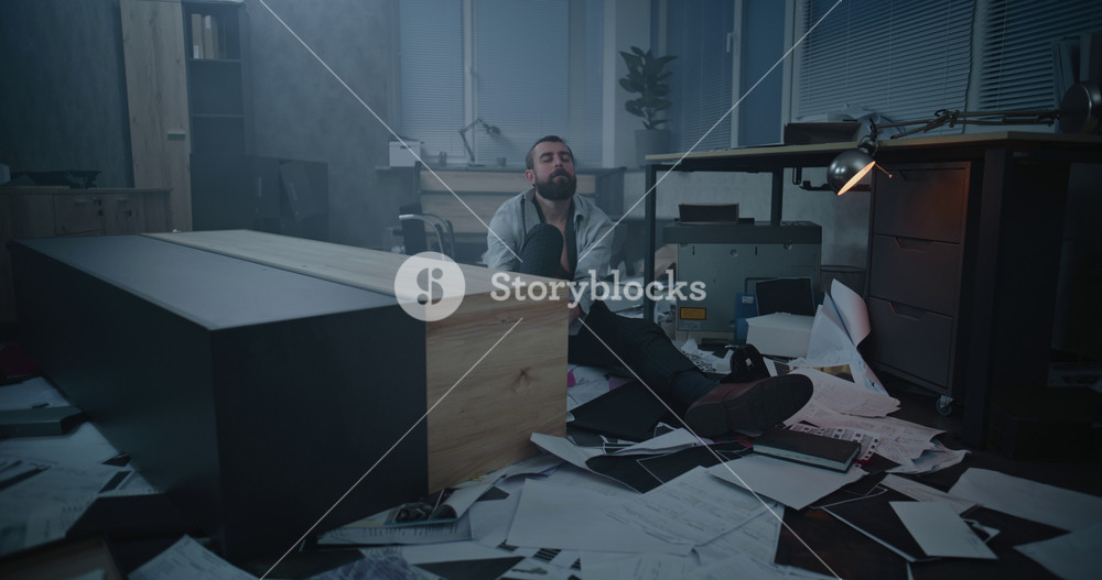 Dolly shot of frustrated fired office worker sitting on floor in destroyed office room. Depressed businessman after fit of rage over