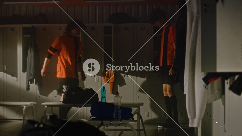 Two Teenage Male Football Players in Orange Uniforms Ball Gently to Each Other Near Lockers in Dark, Sunlit Locker Room. Concept of