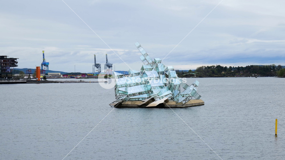 glass and steel iceberg like sculpture floats calm harbor waters cranes waterfront buildings wooded shoreline appear oslo norway floating form modern art