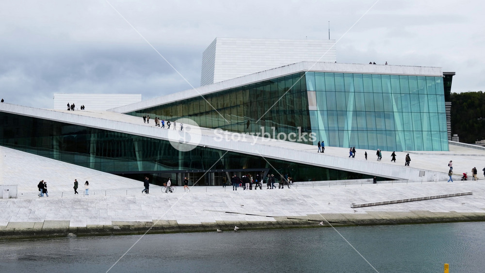 people stroll across gently sloping white marble roof oslo opera house geometric planes oslofjord waters wooded hills city buildings appear modern architecture