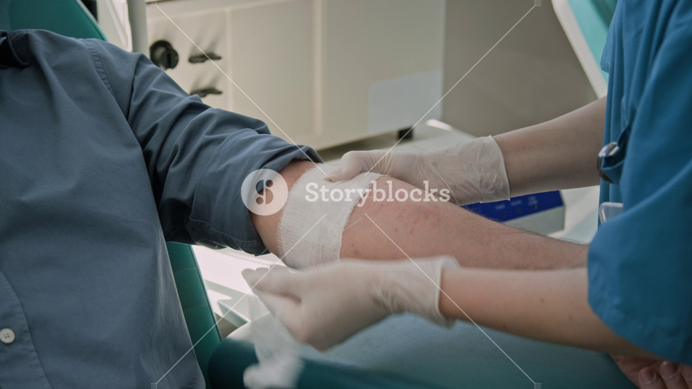 Close-up shot of unrecognizable nurse in gloves and blue medical scrubs bandaging arm of male blood donor in clinic