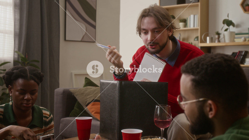 Tilt down shot of group of friends listening to game master behind screen and eating snacks while playing tabletop RPG game at home in cozy living room