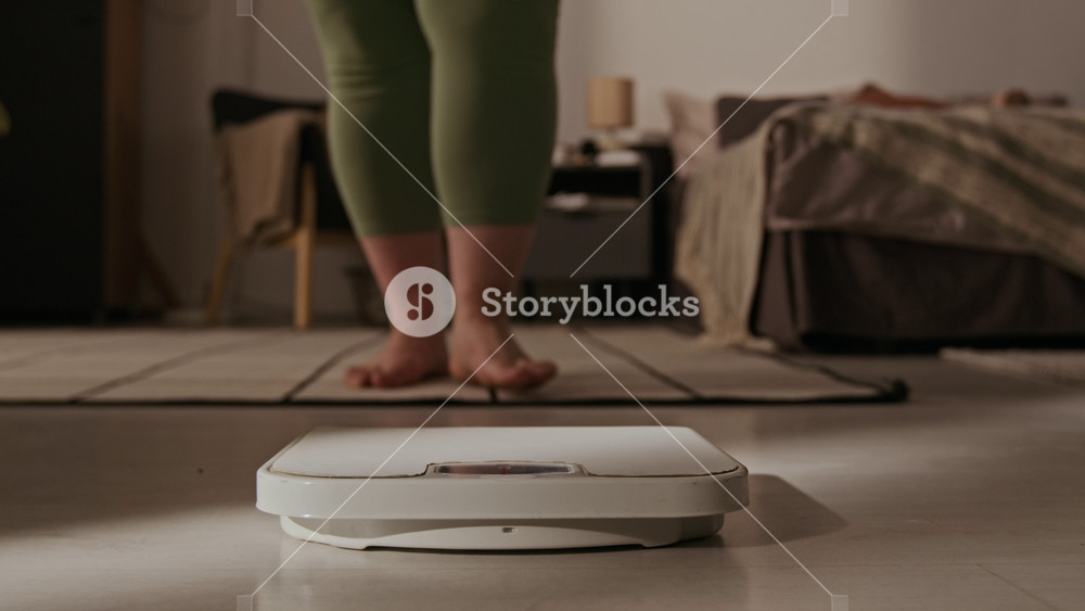 Plus-size Caucasian young adult woman stepping barefoot onto floor scale at home. Clip is perfect for weight loss programs, body positivity campaigns, and fitness tracking apps