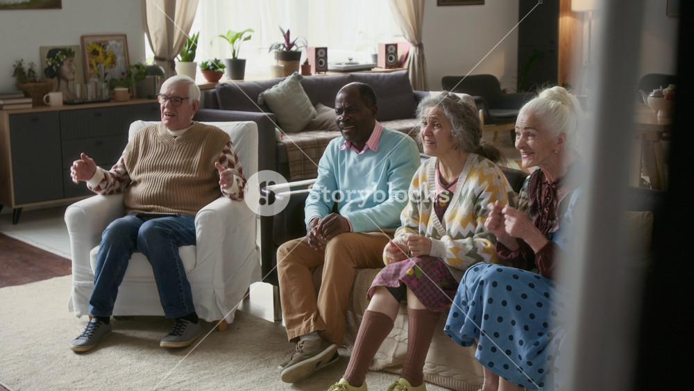 Group of excited elderly friends sitting on sofa, watching sports match on TV, raising their hands and celebrating victory of favourite team in living room of nursing home